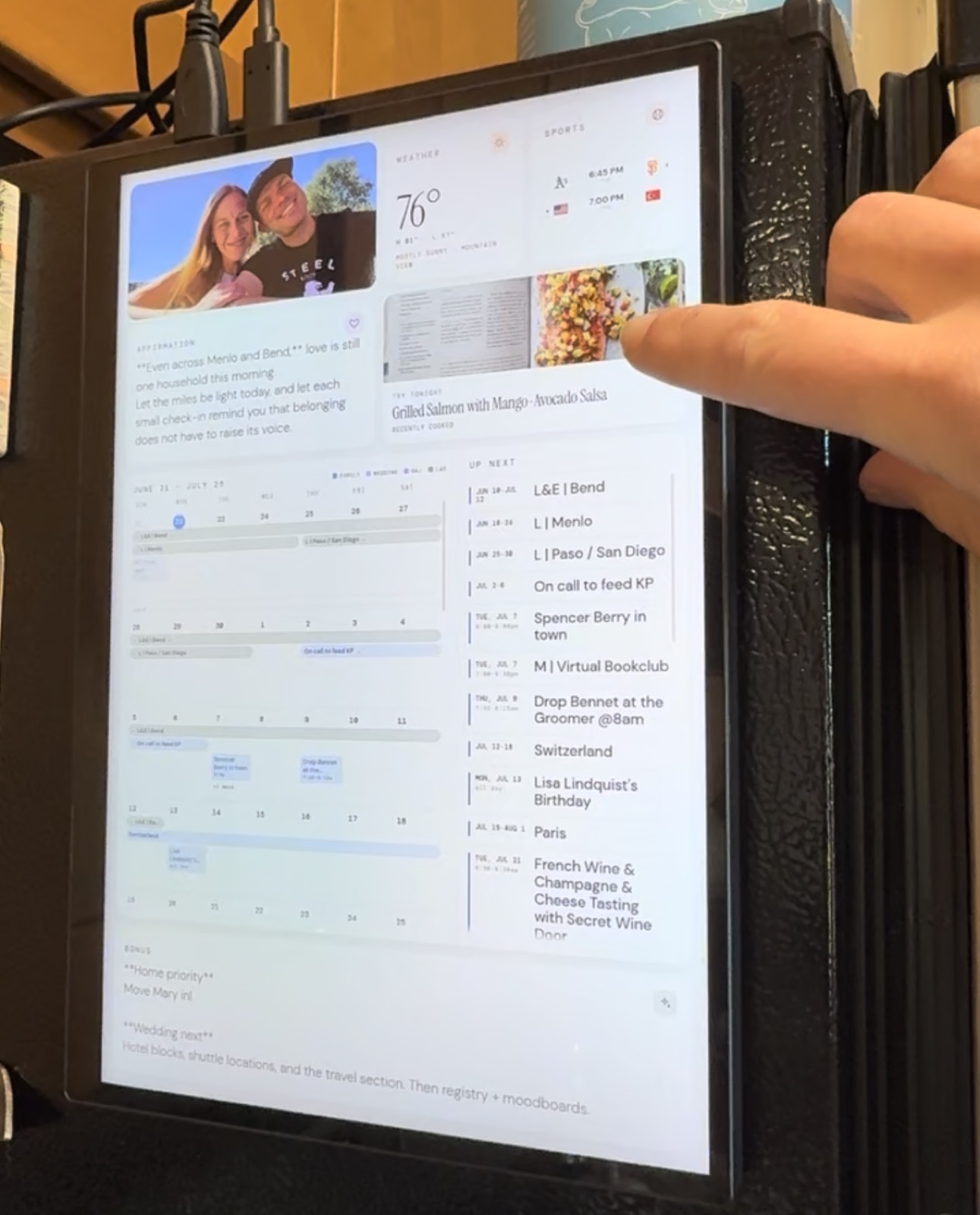 A hand tapping a recipe card on an always-on kitchen tablet showing the week's meal plan, calendar, and weather.